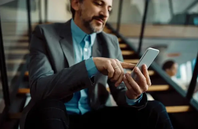 Homem de negócios usando smartphone em ambiente empresarial moderno, vestindo terno cinza e camisa azul, focado na tela.