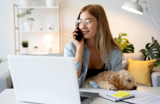 Mulher jovem com óculos, sorrindo ao falar ao telefone, sentada em um ambiente bem decorado com um laptop, cachorro de estimação ao lado, passando uma sensação de conforto e produtividade.