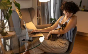 Uma mulher com cabelos encaracolados e óculos trabalha em um laptop em uma mesa de vidro, com um cachorro pequeno e escuro aninhado em seu colo.